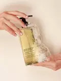 Hands holding a bottle of Shu Uemura Gentle Radiance Deep Cleanser weightless shampoo with foam and bubbles surrounding the bottle.