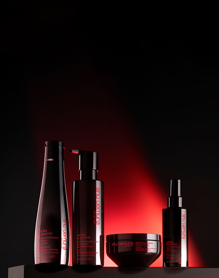 Four black bottles of Shu Uemura Ashita Supreme intense revitalization haircare products, including shampoo, conditioner, mask, and spray, displayed on a black and red gradient background.