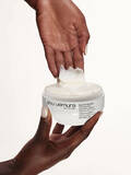 Two hands holding an open jar of Shu Uemura Izumi Tonic Strengthening Treatment with a dollop of cream being lifted.