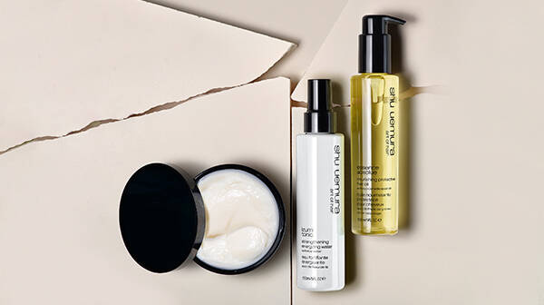 Three Shu Uemura haircare products: a jar of cream, a white bottle, and a yellow bottle, arranged on a beige cracked surface.