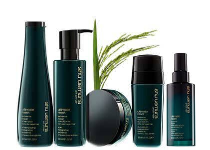 A group of Shu Uemura Ultimate Reset hair care products, including shampoo, conditioner, mask, serum, and spray, with green packaging and a plant in the background.