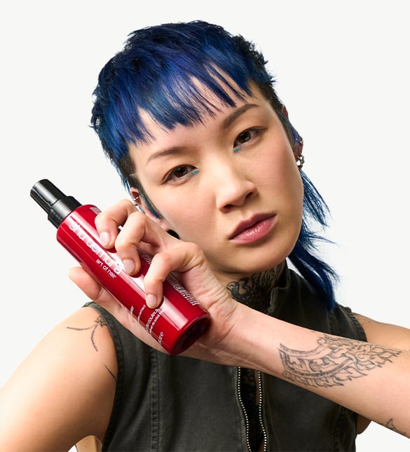 A person holds a red bottle of Shu Uemura Blow Dry Oil Spray, showcasing a tattooed arm.