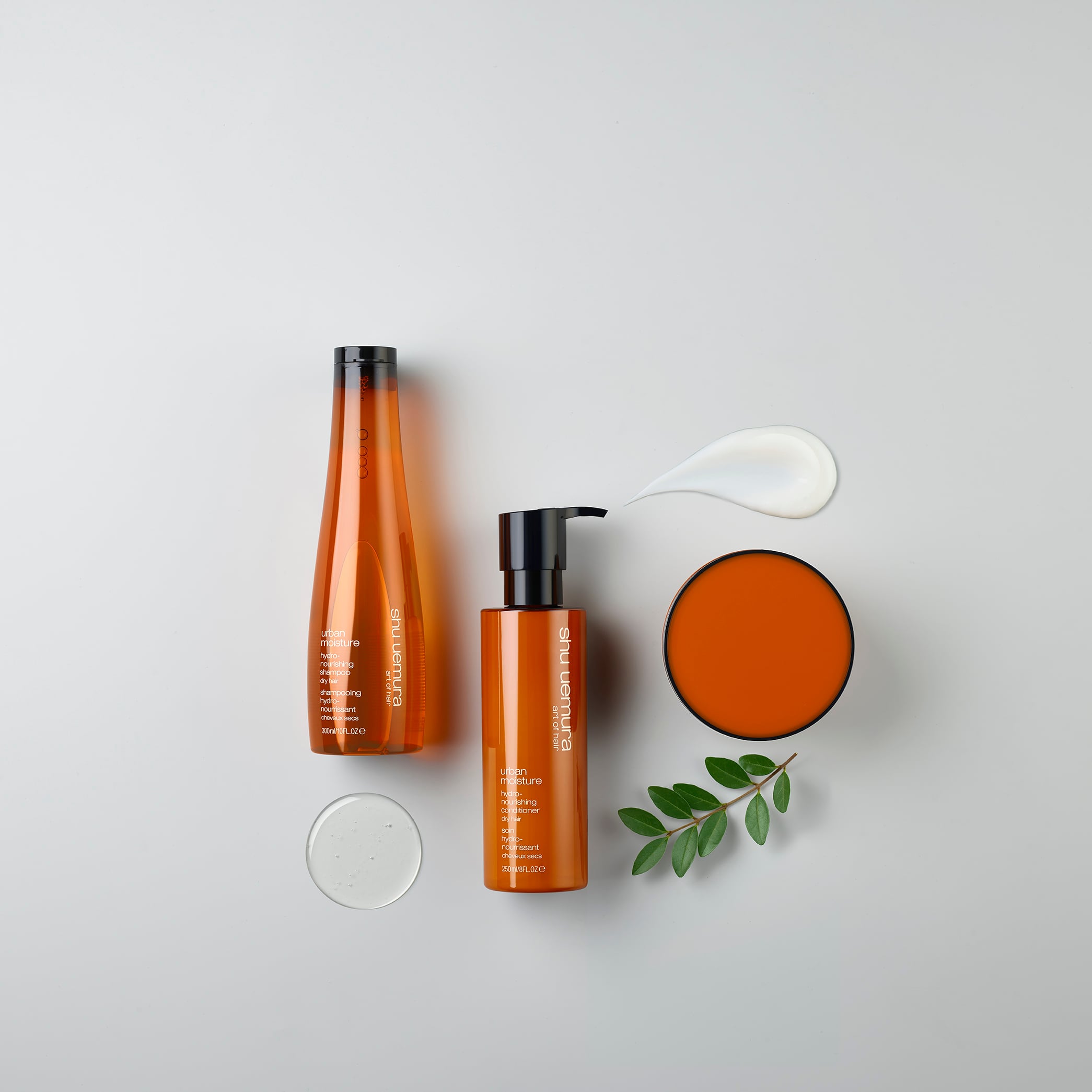 A collection of Shu Uemura Urban Moisture haircare products, including a shampoo, conditioner, mask, and spoon, displayed on a white surface.