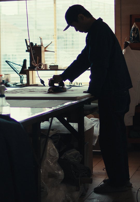 A person ironing fabric on a large table in a workshop filled with sewing tools and materials.
