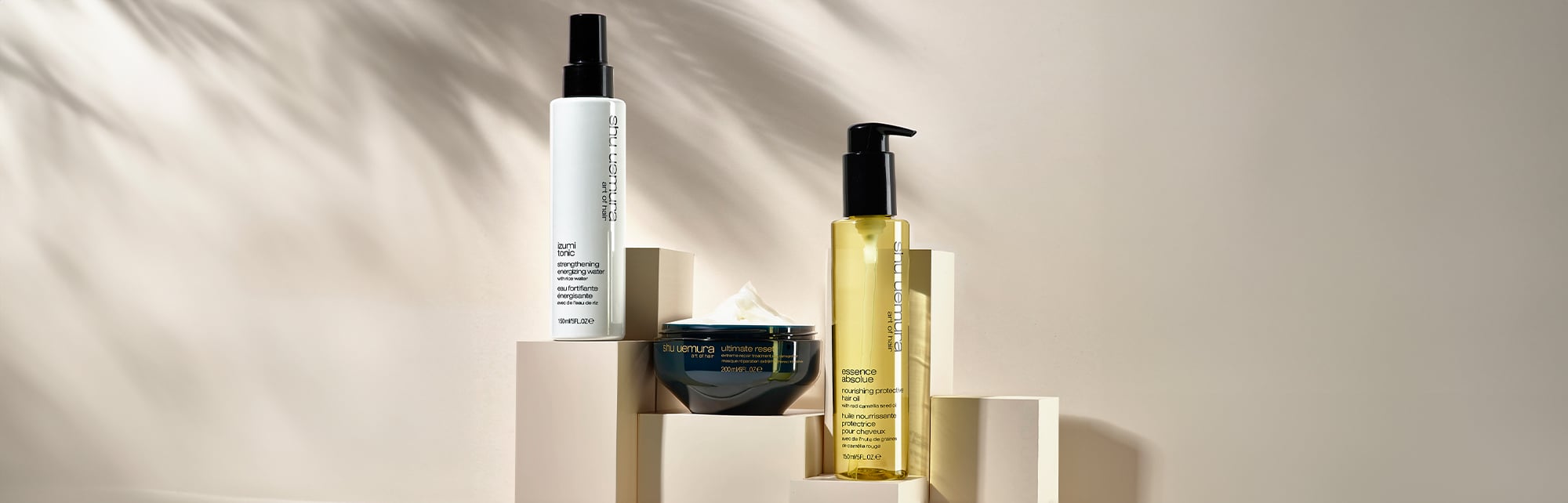 Shu Uemura Izumi Tonic strengthening energizing water, Ultimate Reset hair mask, and Essence Absolue nourishing protective hair oil displayed on beige blocks with soft shadows.